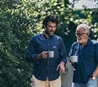 Older man in blue shirt talking to younger man in checkered shirt outside holding mugs