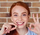 Woman with red hair smiling holding extracted tooth and making “ok” symbol with her hand